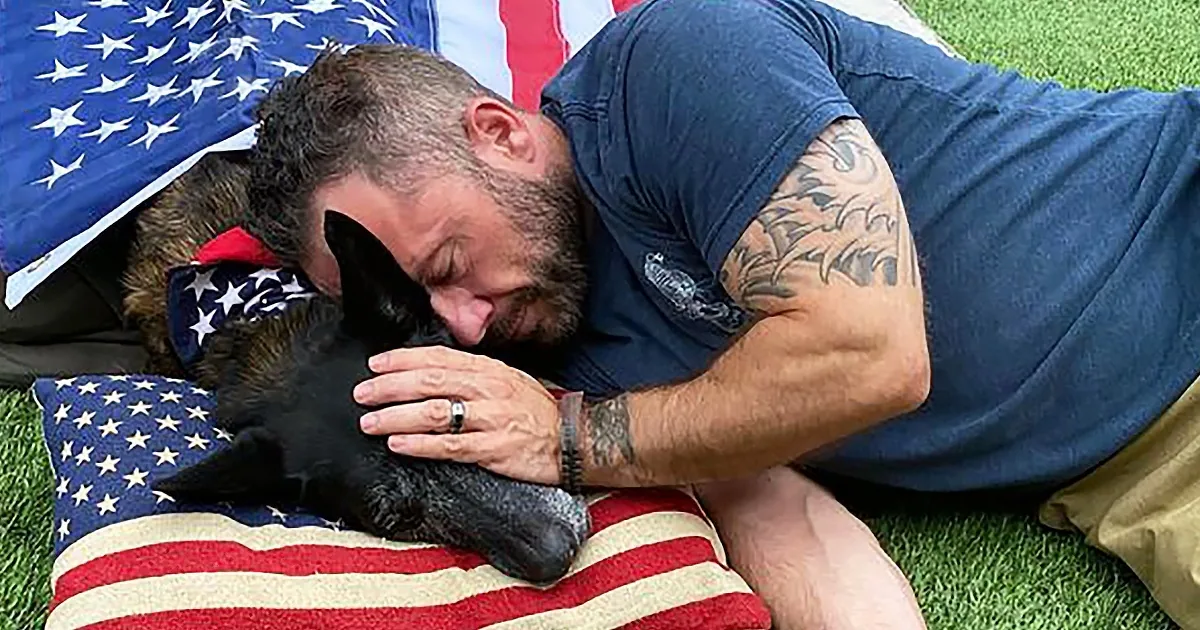 A man with tattoos hugs a black dog on an American flag pillow.