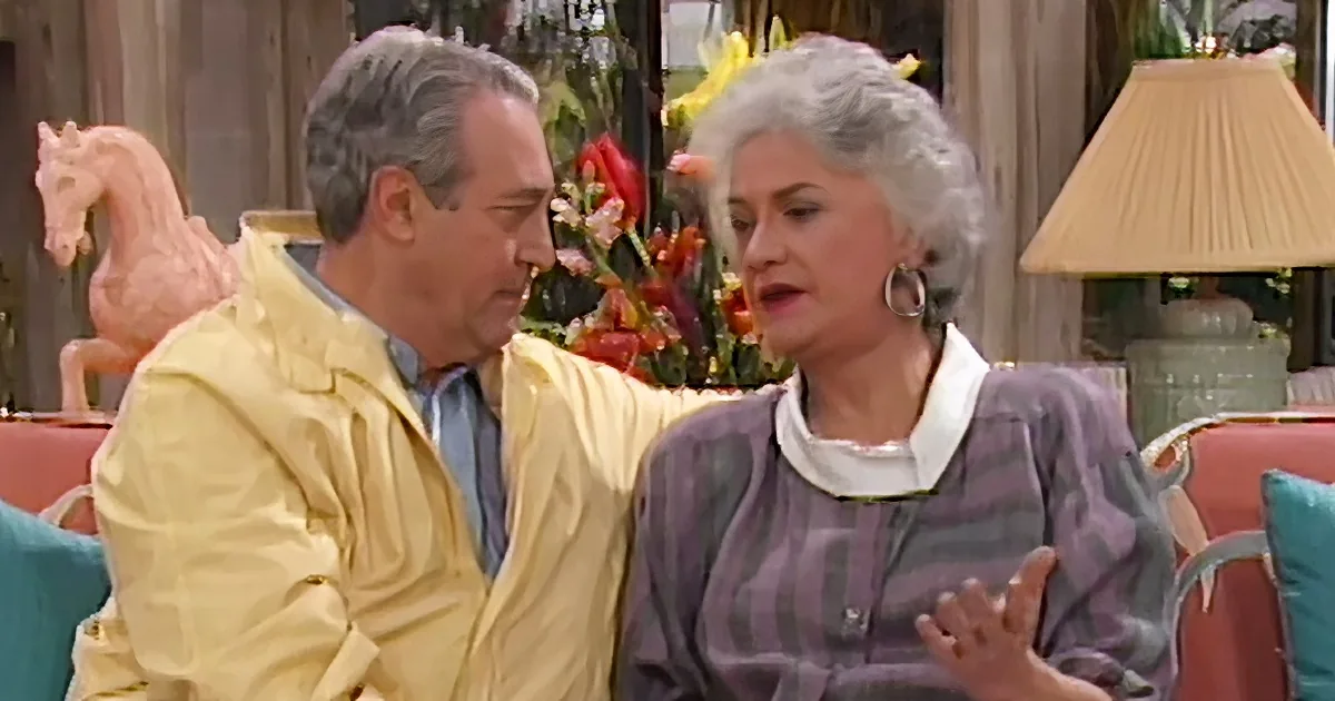 A man in a yellow jacket is talking to a woman in a purple dress, both seated in a living room setting.