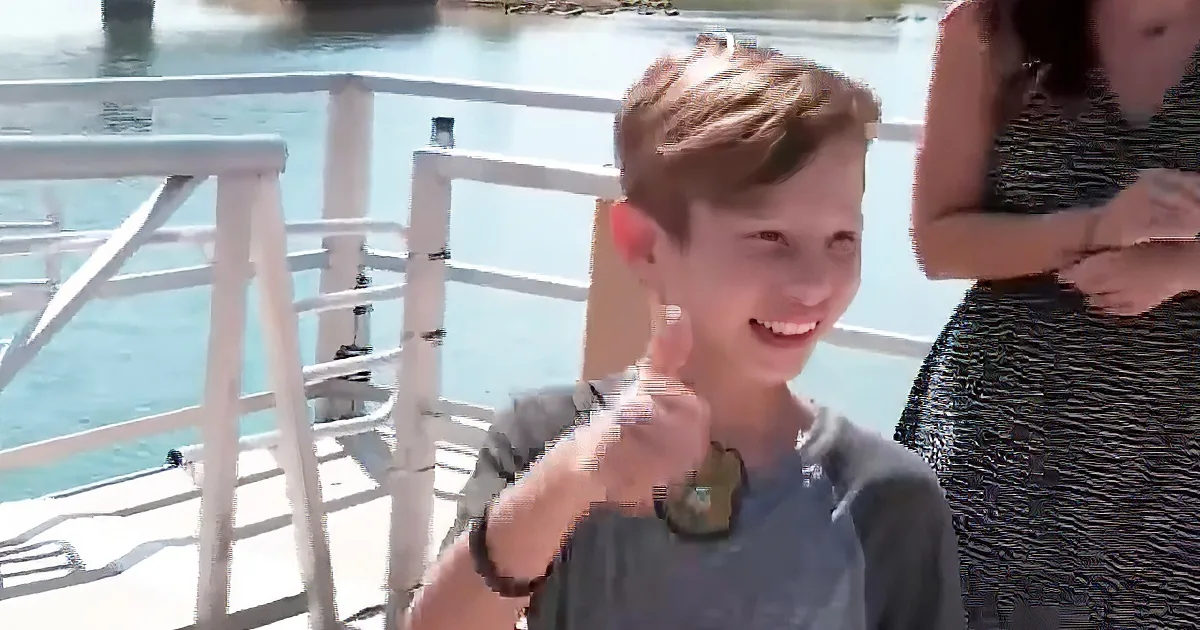 A boy giving a thumbs-up on a boat, smiling, with a woman in the background.