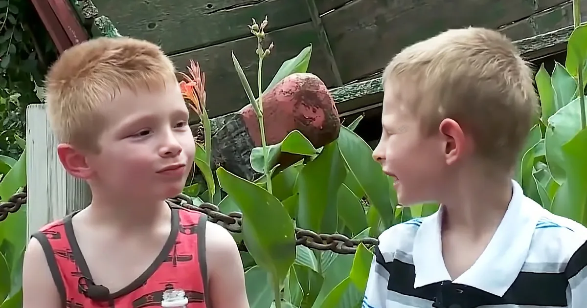 Two young boys, one in red tank top, the other in striped shirt, stand facing each other in a lush garden setting.