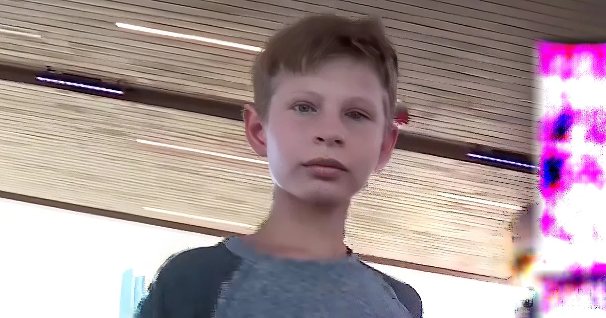 A young boy with short brown hair and a gray shirt stands in front of a window with horizontal blinds.