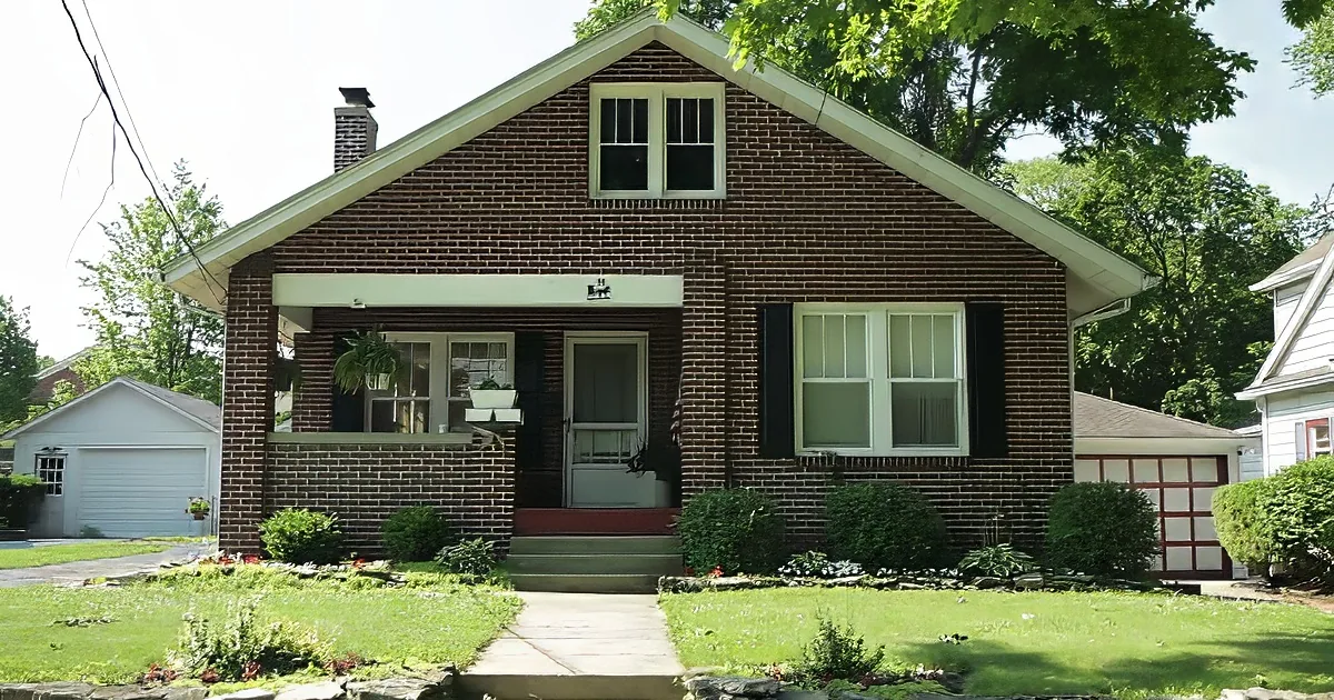 A charming brick house with white trim, a porch, and a small garden, surrounded by trees and a clear sky.
