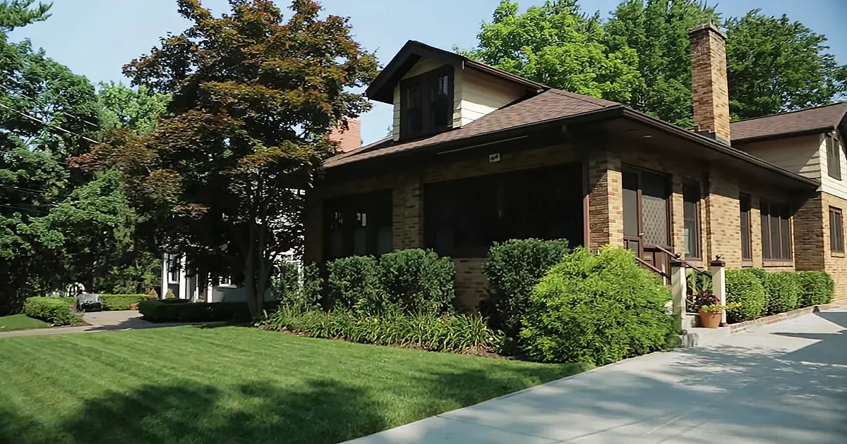 A charming house with a sloped roof, brick chimney, and lush green lawn surrounded by trees and bushes.