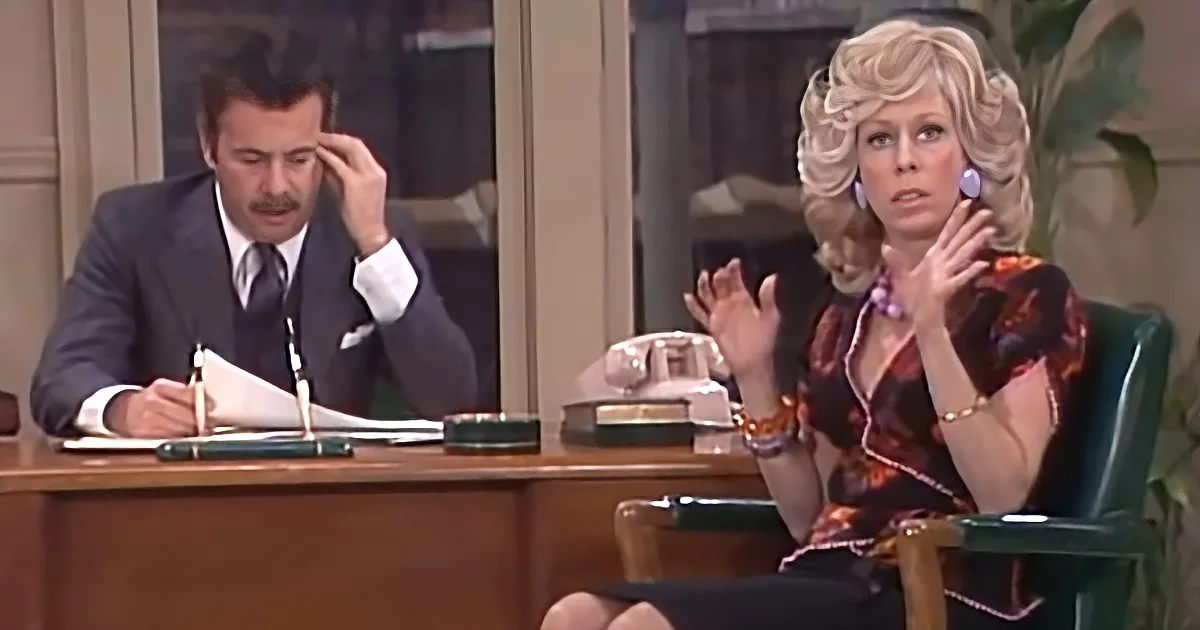A man in a suit writes at a desk while a woman in a floral dress gestures with her hands.