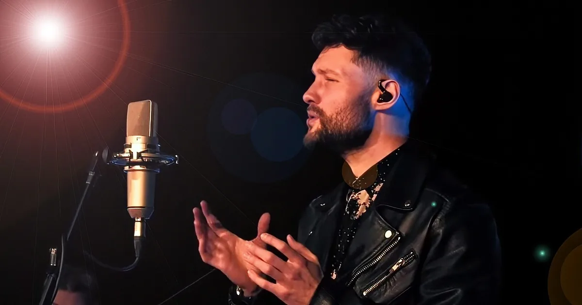 A man in a leather jacket sings into a microphone, hands clasped, with a bright light in the background.