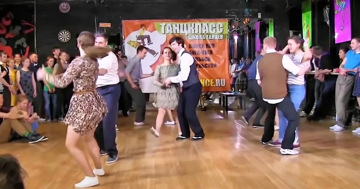A couple in a dance class, the woman in a patterned dress, the man in a white shirt and vest, surrounded by other dancers...