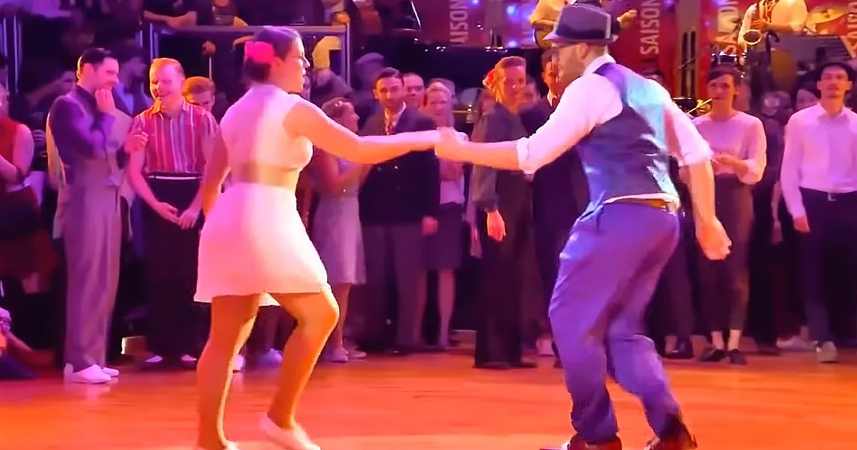 A couple in vintage attire dances on a wooden floor, surrounded by an audience in a lively setting.