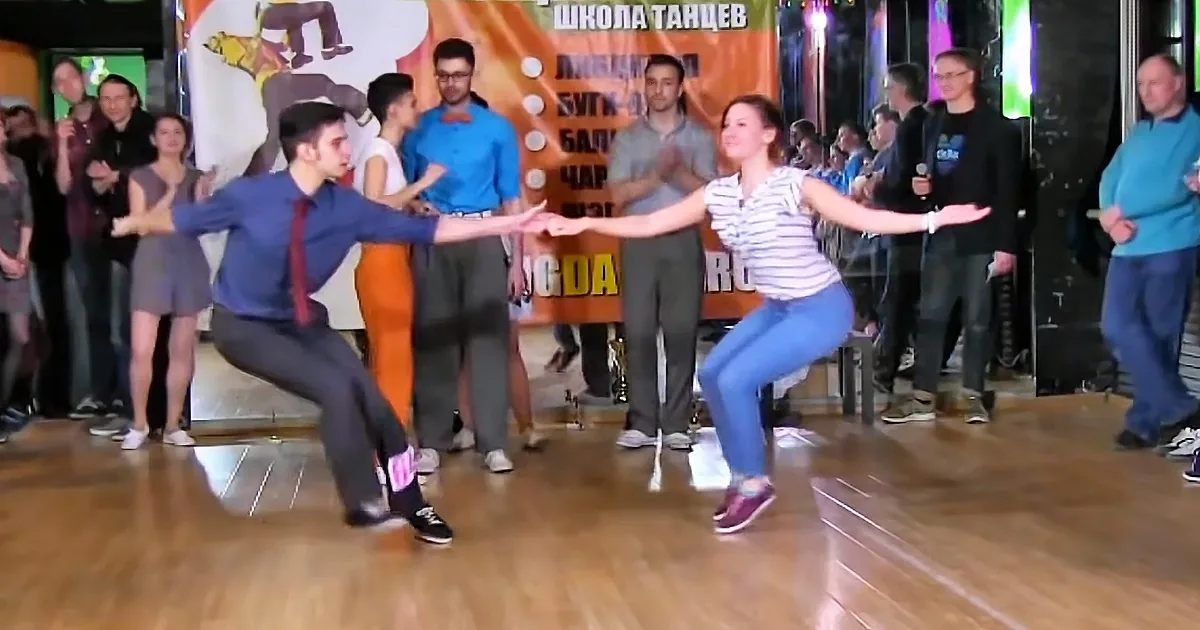 A man and woman dance in a studio, holding hands, with spectators watching in the background.