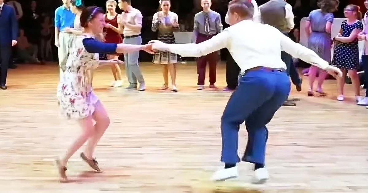 A man and woman are dancing together on a wooden floor, surrounded by onlookers.