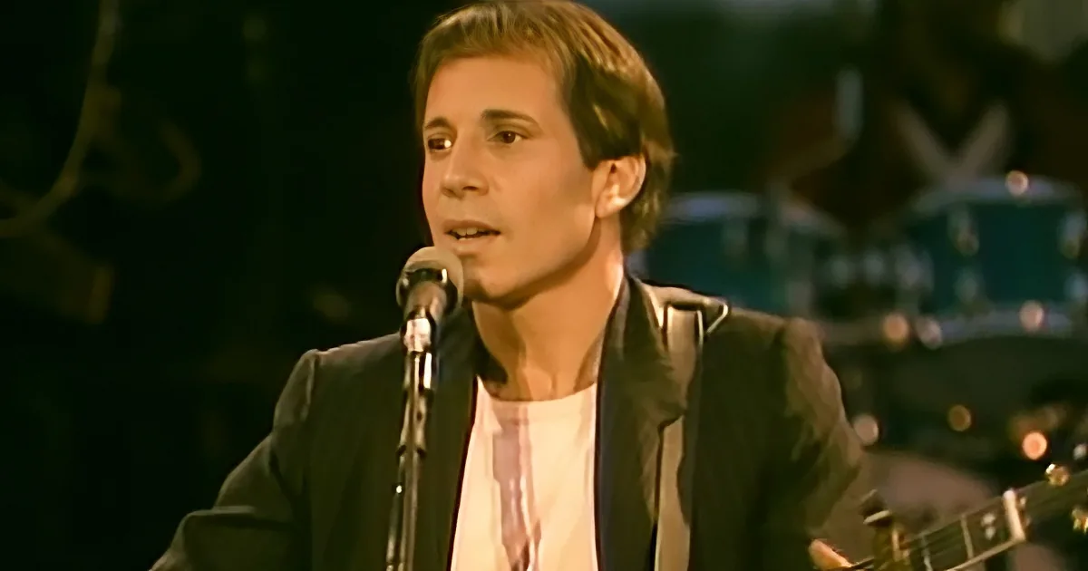 A man in a black jacket sings into a microphone, with a drum set in the background.