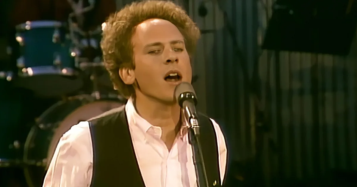 A man with curly hair sings into a microphone, wearing a white shirt and black vest, with a drum set in the background.