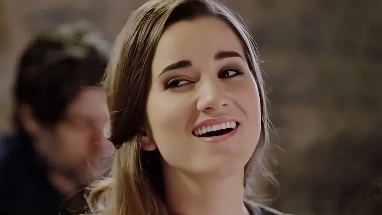 A woman with long brown hair smiles widely, her eyes looking to the side, while a blurred man sits behind her.