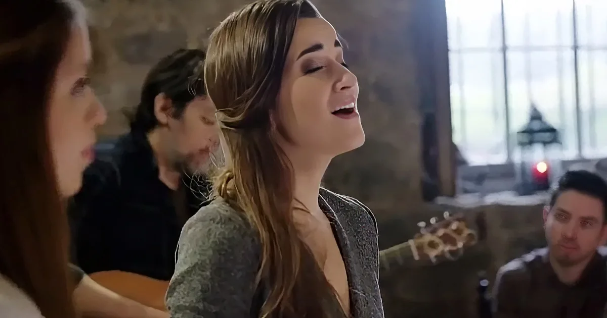 A woman sings passionately in a dimly lit room with a guitarist and another person in the background.