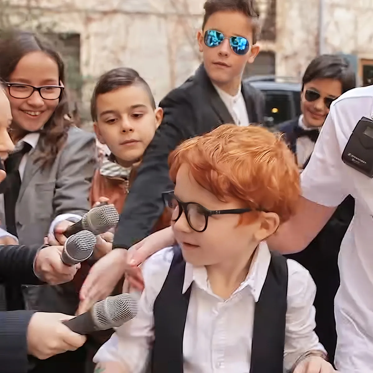 A young boy with red hair and glasses is surrounded by people in suits, holding microphones, in an outdoor setting.