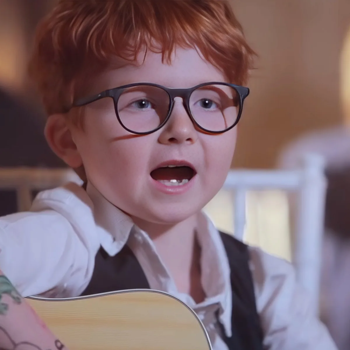 A young boy with red hair and glasses is singing while holding a guitar.