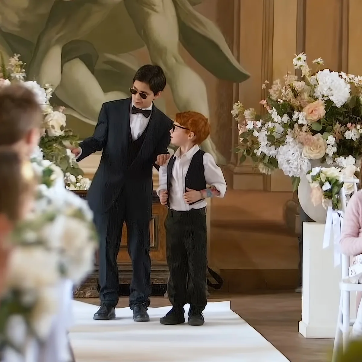 A young boy in a suit and sunglasses stands on a white carpet, holding hands with another boy in a vest and glasses.