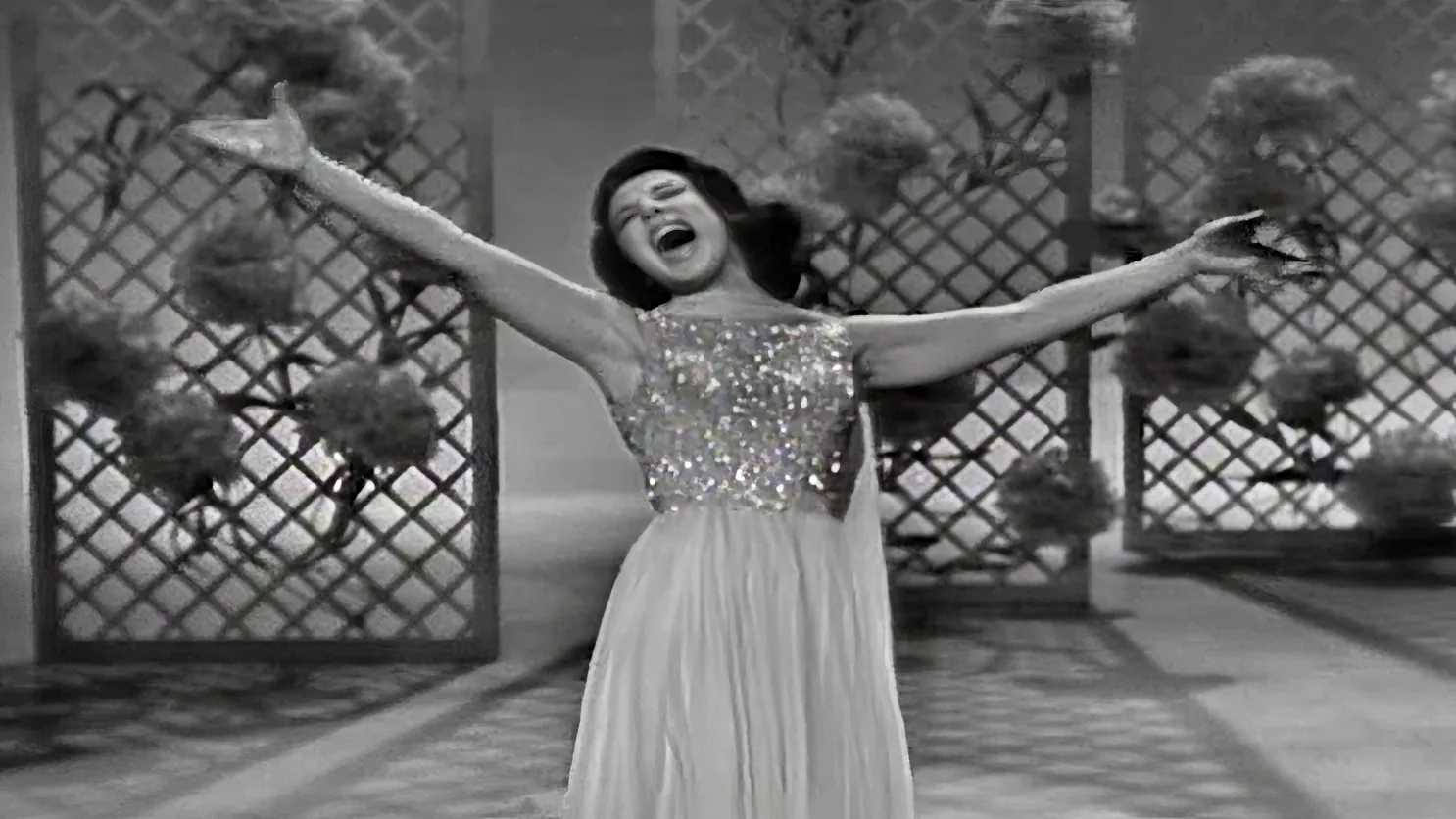 A woman in a sparkling dress and hat is joyfully dancing with arms outstretched.