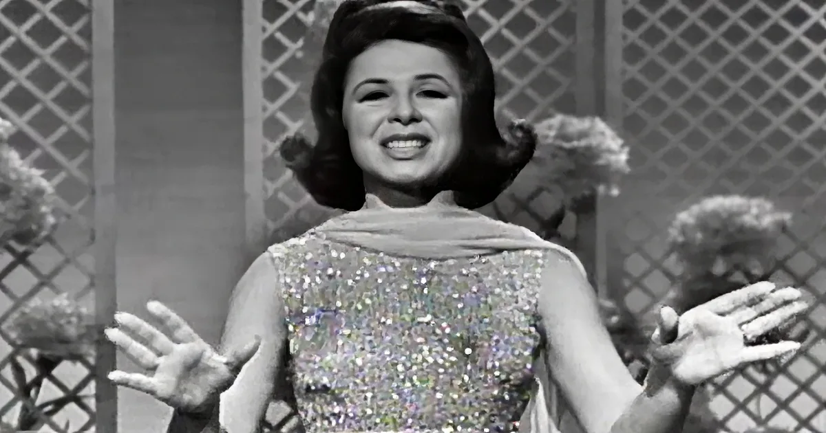 A woman in a sparkly dress sings with her hands raised, surrounded by lattice and flowers.