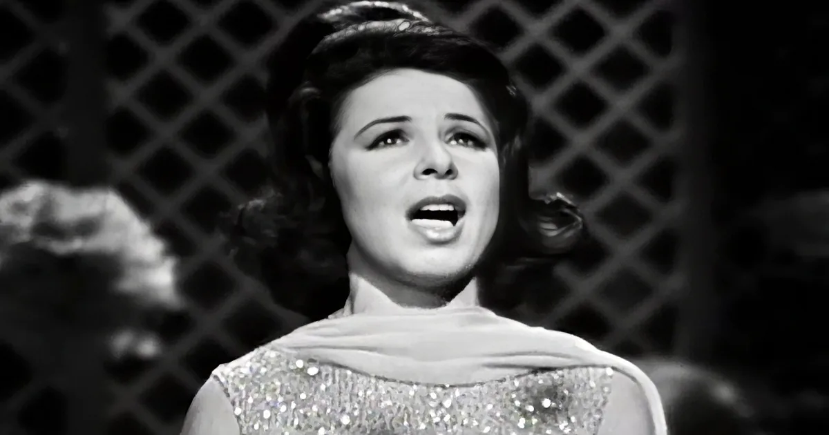 A woman in a sparkly dress sings passionately, her mouth open, with a lattice background.