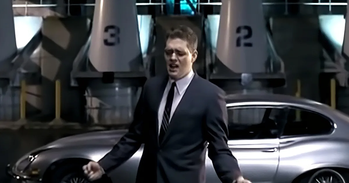 A man in a suit stands beside a sleek car, with numbered structures in the background.