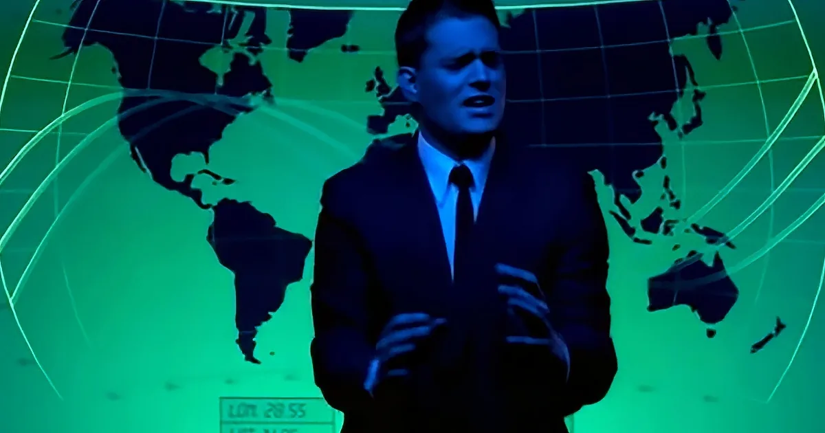 A man in a suit stands in front of a green world map backdrop, gesturing with his hands.