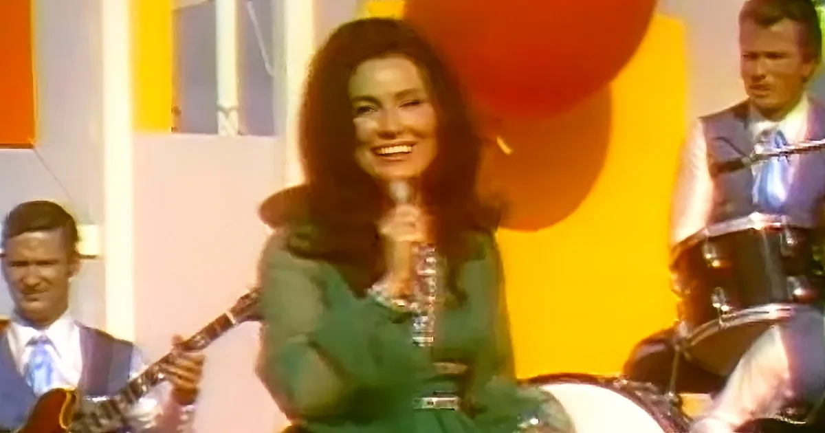 A woman in a green dress smiles while holding a microphone, with a band and colorful background behind her.