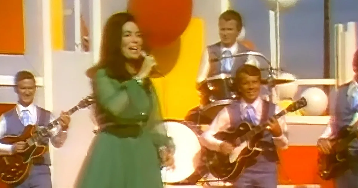 A woman in a green dress smiles on stage with a band playing guitars and drums.