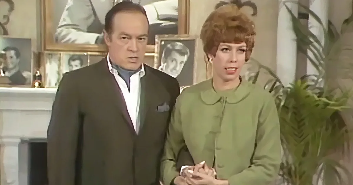 A man in a dark suit and a woman in a green dress stand together, surrounded by framed pictures and a plant.