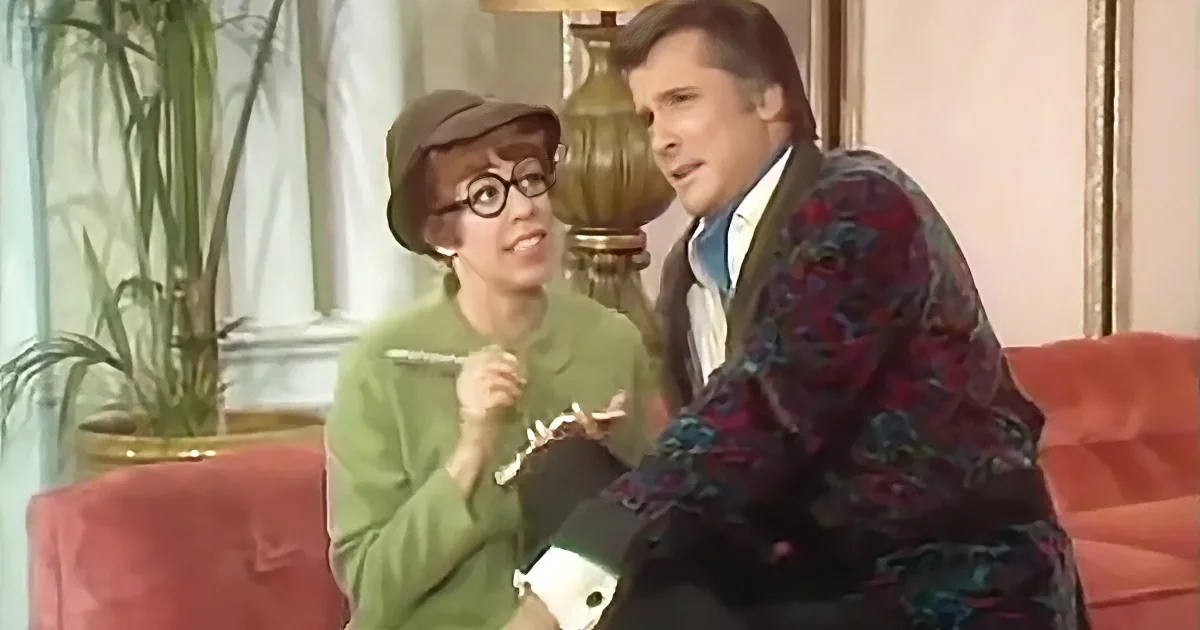 A man in a patterned suit sits beside a woman in a green outfit on a red couch, both engaged in conversation.