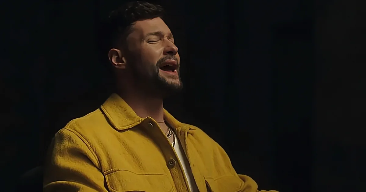 A man in a yellow jacket sings passionately with his eyes closed, illuminated by a spotlight.