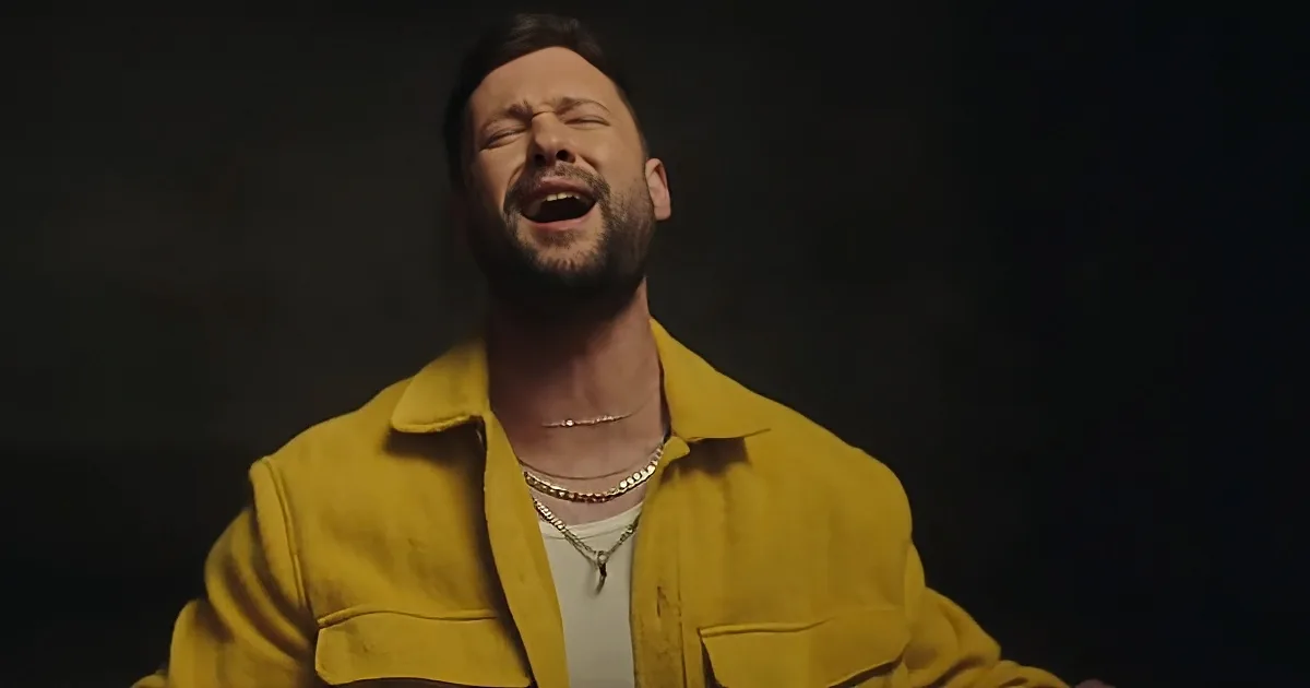 A man in a yellow jacket and white shirt is singing with his eyes closed, wearing a necklace.