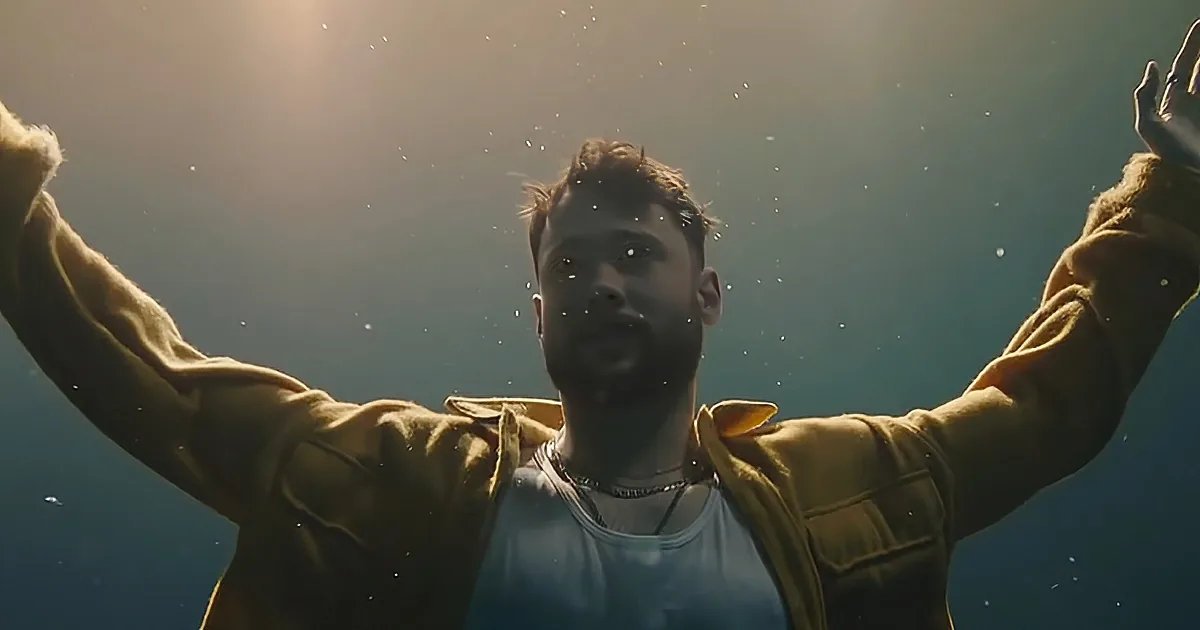 A person in a yellow jacket and white tank top, arms outstretched, appears to be underwater with light filtering through.