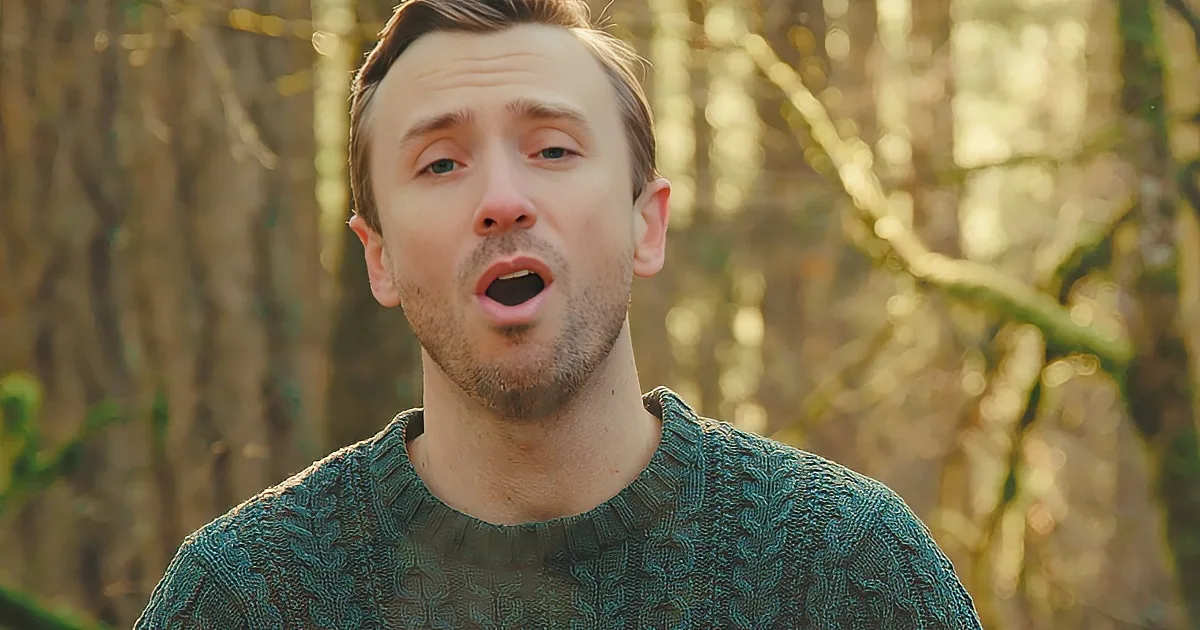 A man in a green sweater stands in a forest, mouth slightly open, with trees blurred in the background.