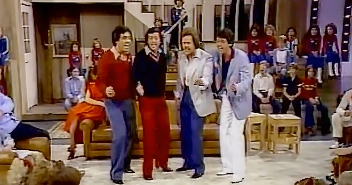 Four men in colorful 70s attire are dancing in a living room with a crowd watching.