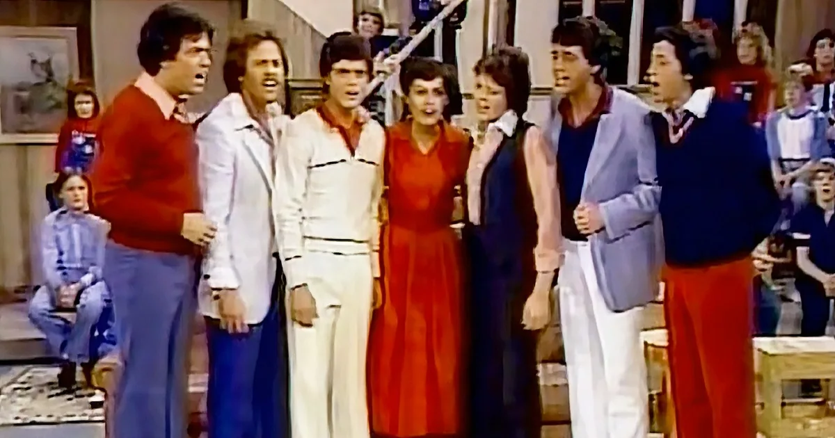 A group of people in 1970s attire sing together on a stage, with an audience in the background.
