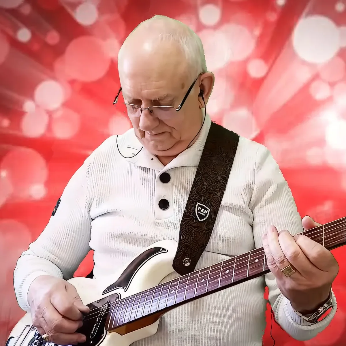 An elderly man with glasses plays a white guitar against a red bokeh background.