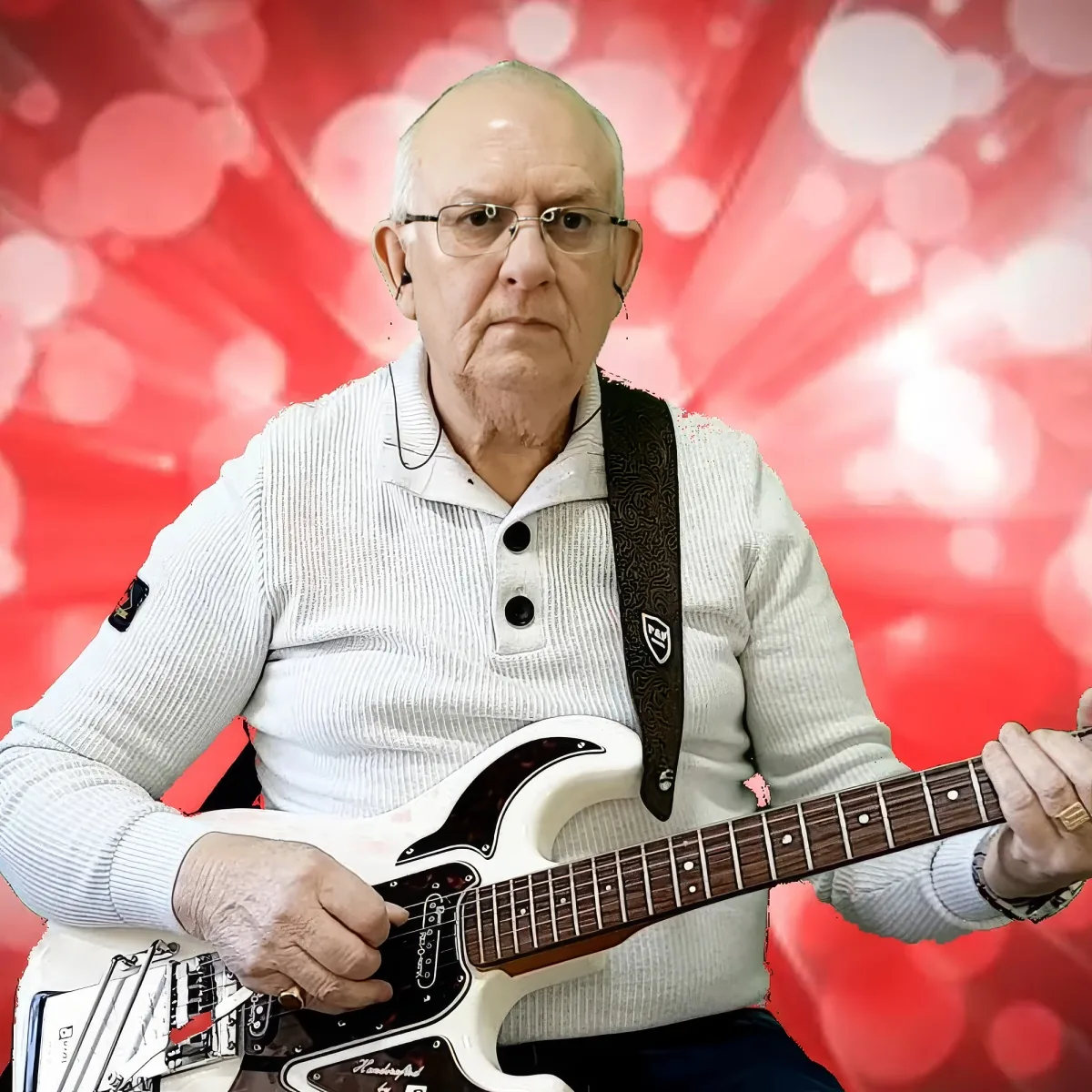 An older man wearing glasses plays a white electric guitar against a red, bokeh background.