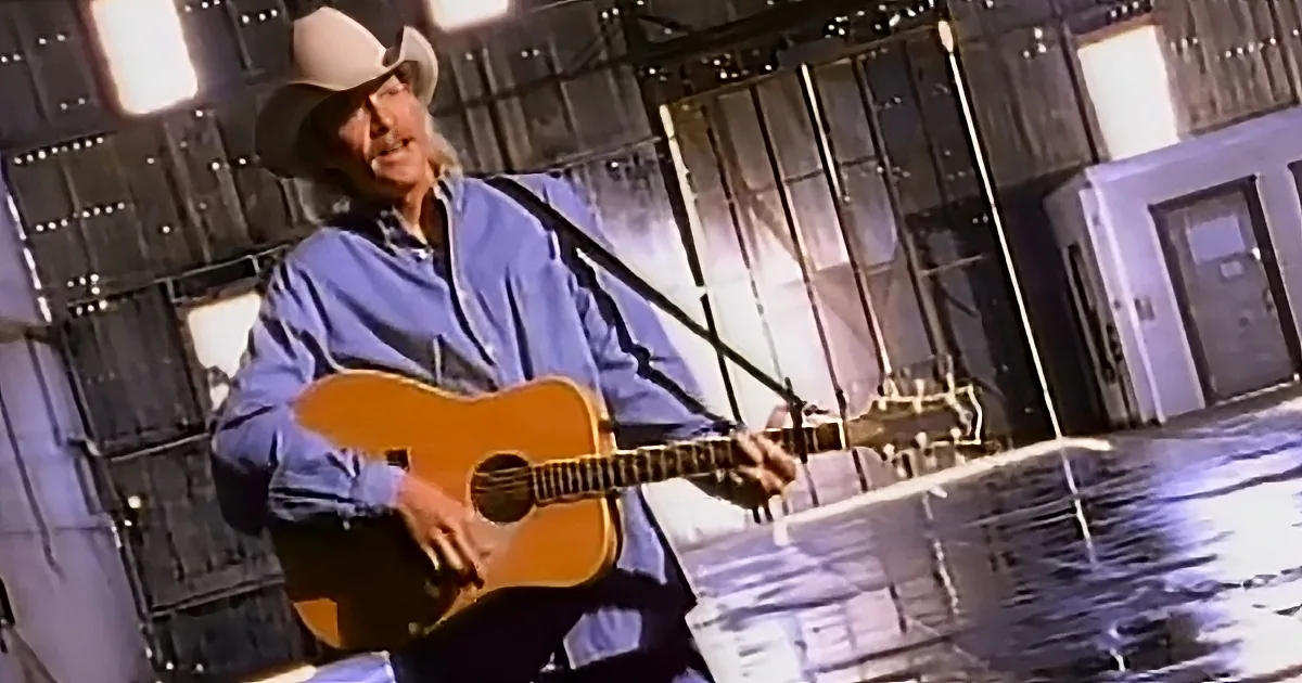 A man in a cowboy hat plays an acoustic guitar on stage with a microphone and lights.