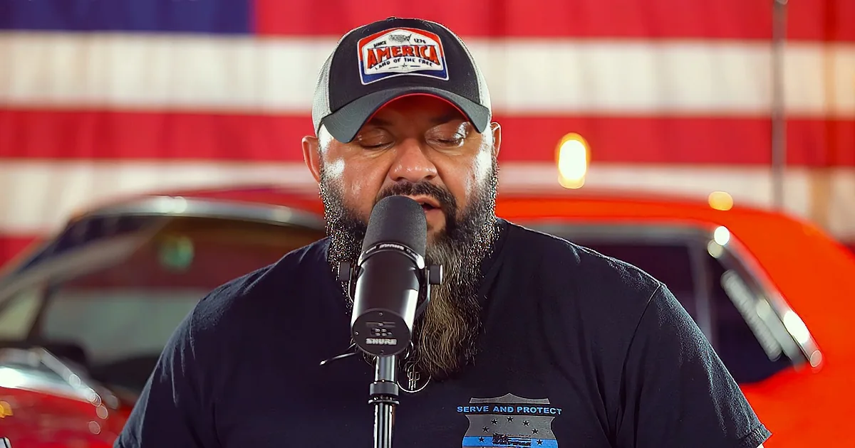 A man with a beard and a "America" hat sings into a microphone in front of an American flag.