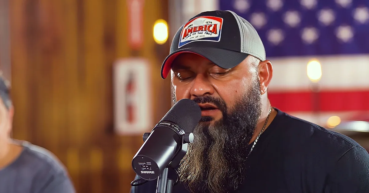 A man with a beard and a "America" cap speaks into a microphone in front of an American flag.