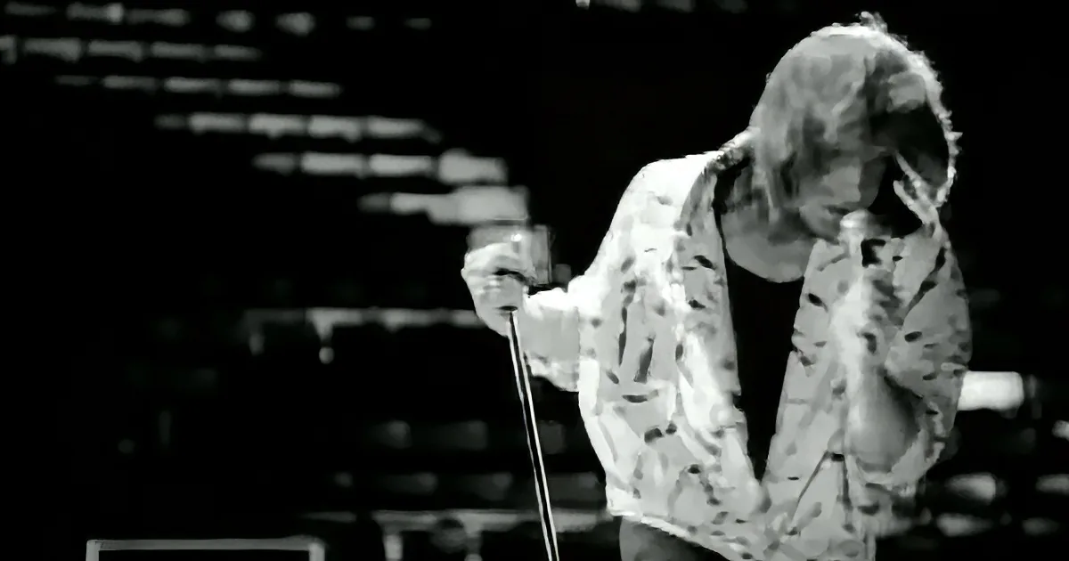 A person in a patterned jacket sings into a microphone, with a blurred background and text.