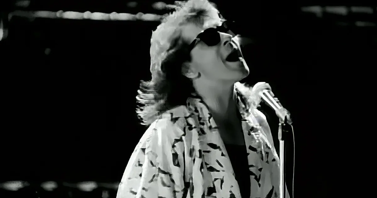 A person wearing sunglasses and a patterned shirt sings into a microphone on stage.