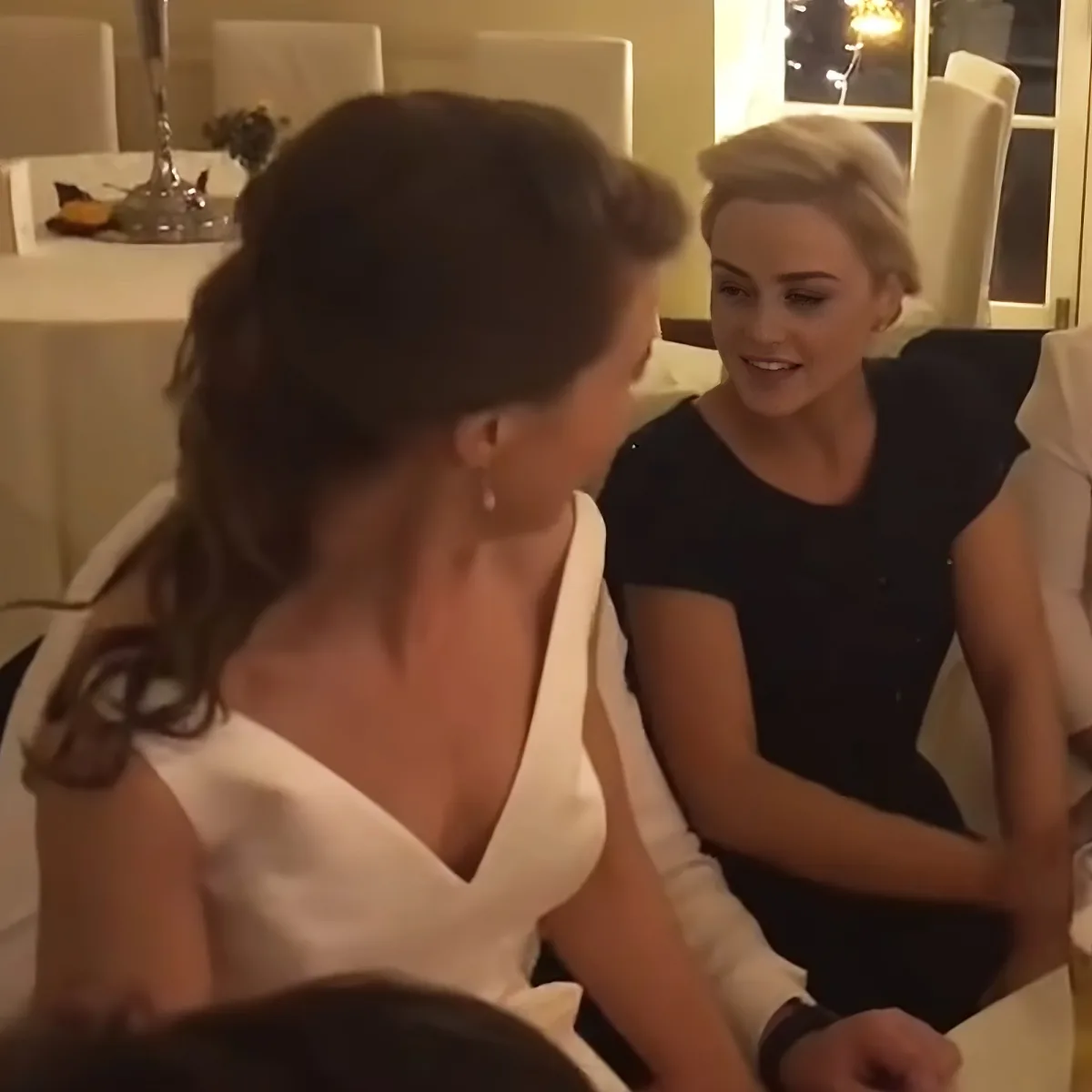 A woman in a white dress is seated, facing another woman in a black dress who is smiling.
