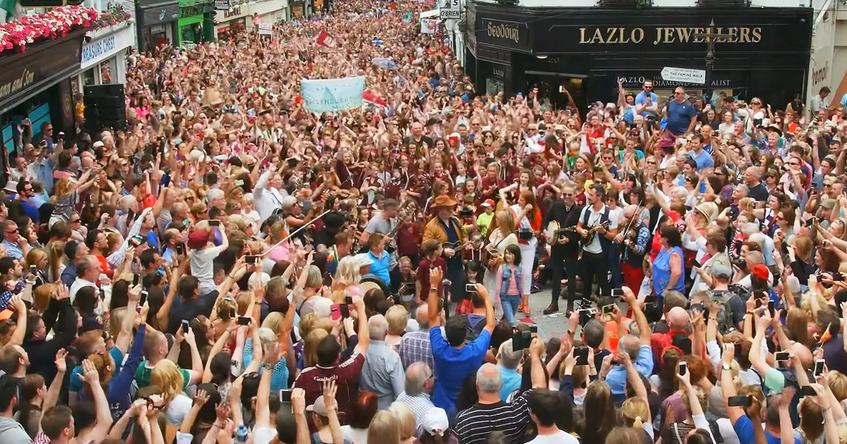 A jubilant crowd celebrates with musicians and cameras in a lively street scene.