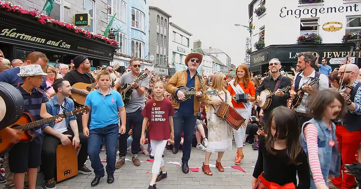 A lively street performance with musicians and a crowd, featuring a man in a brown jacket playing a mandolin.