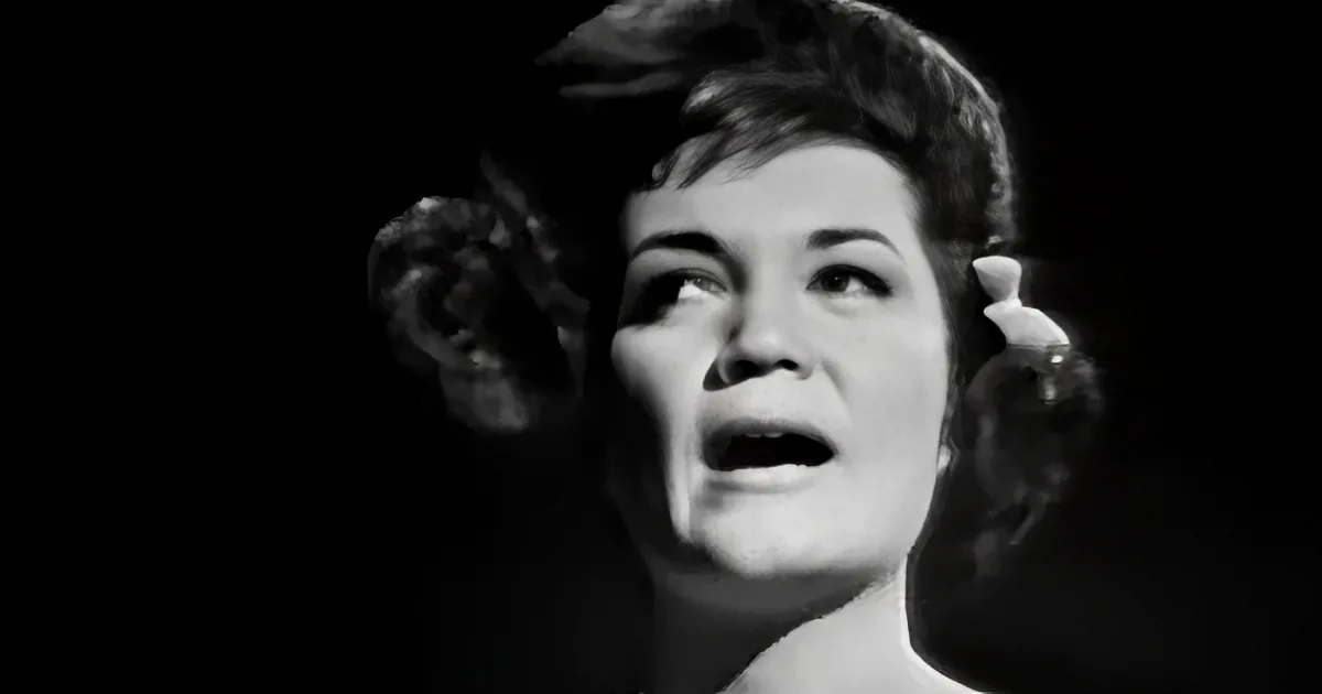A woman with short hair and floral earrings is singing, her mouth open, against a dark background.