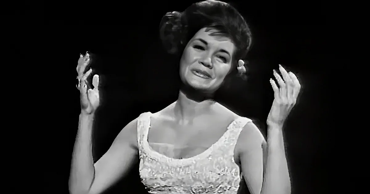 A woman in a white dress with lace details is smiling and clapping her hands.