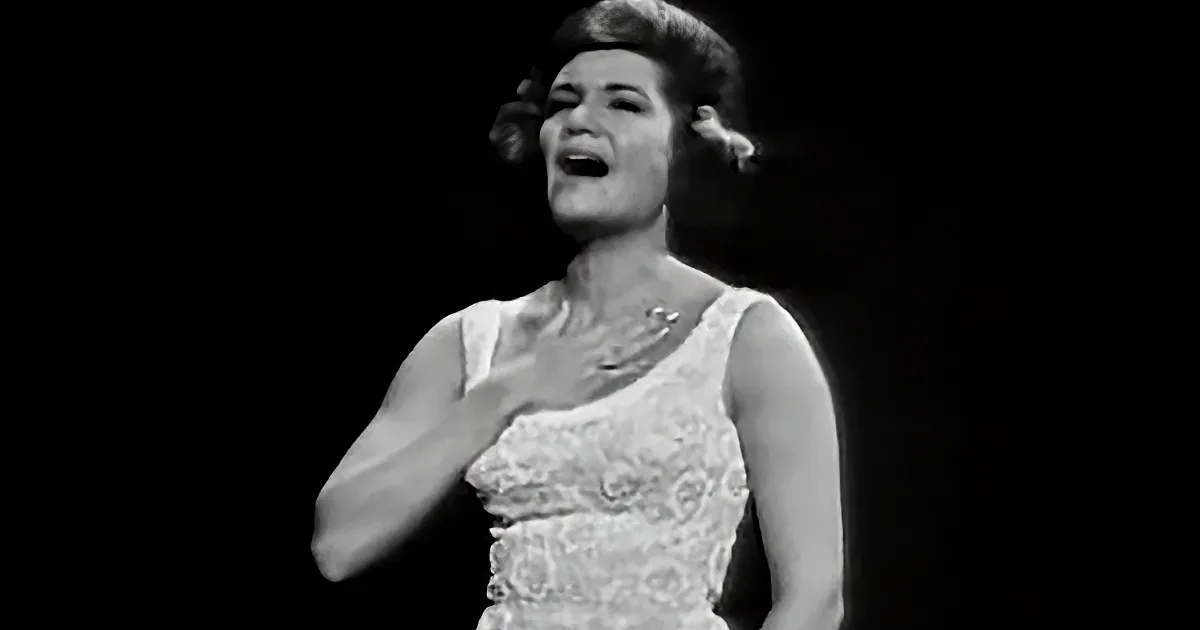 A woman in a white dress is singing passionately with her hand on her chest.