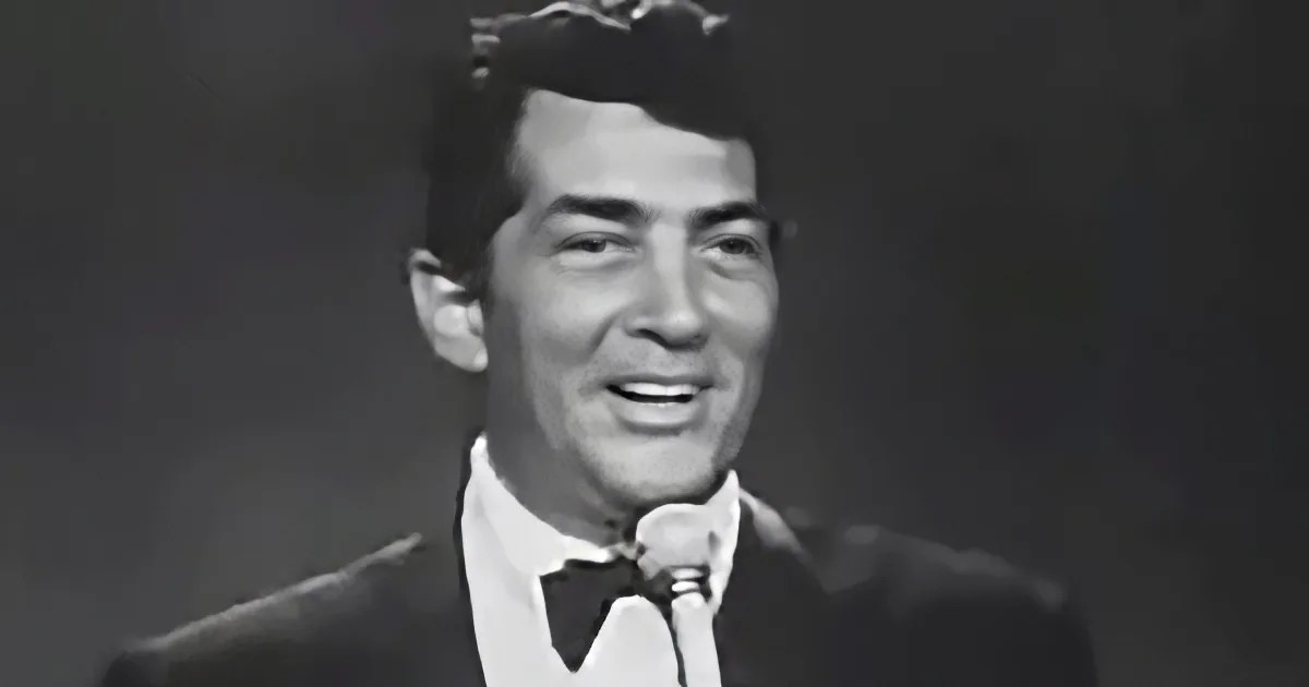 A man in a suit and bow tie smiles into a microphone, likely performing or speaking.