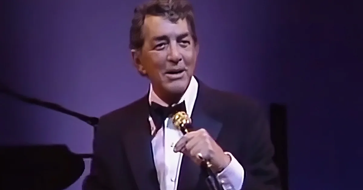 A man in a tuxedo holding a microphone, smiling, and wearing a ring.